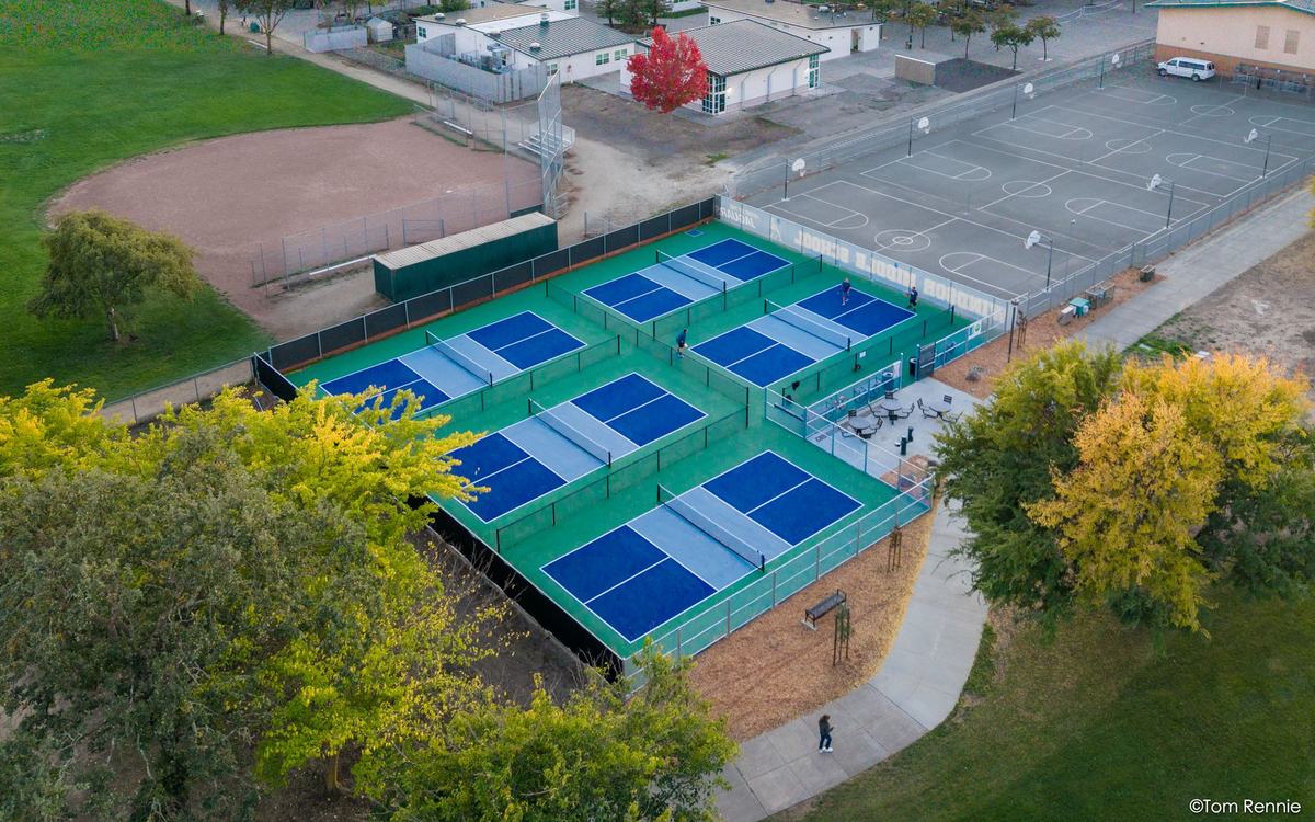 Aerial view at sunset of the five blue and green pickleball courts at Hiram Lewis Park, Windsor, with players on court and the Russian River valley beyond